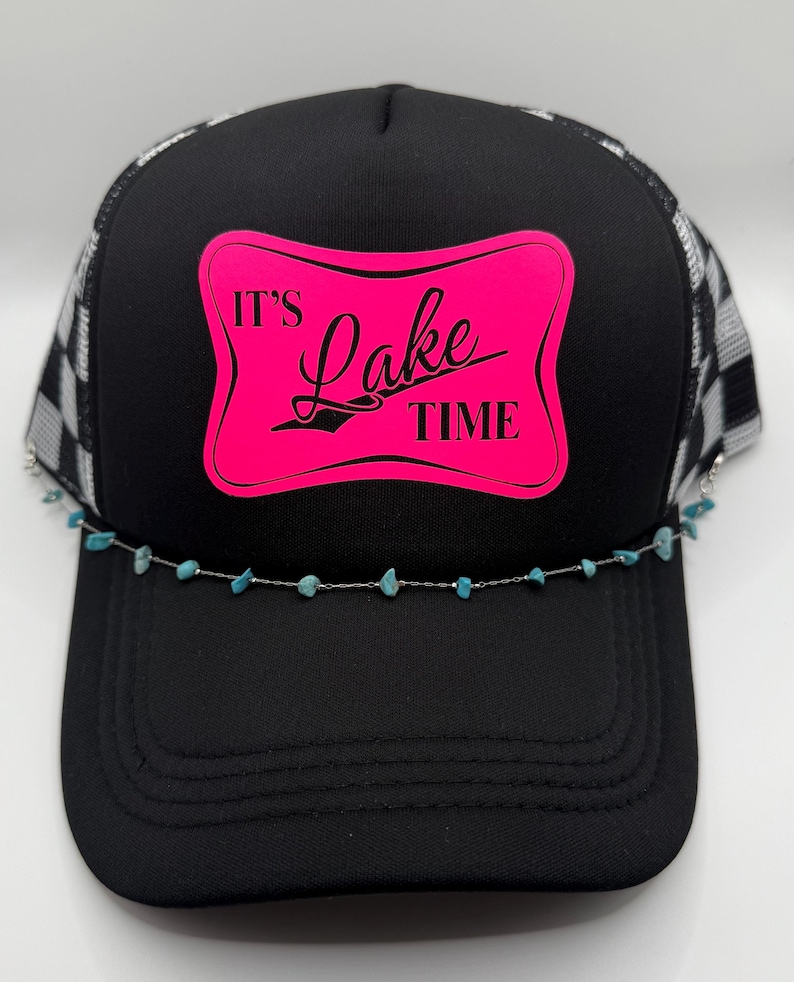 May include: Black trucker hat with a checkered pattern on the mesh and a pink rectangle with the words "It's Lake Time". A silver chain with turquoise beads is attached to the brim.