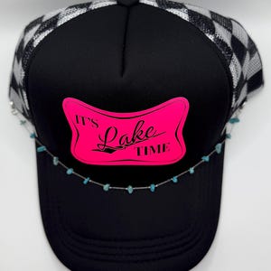 May include: Black trucker hat with a checkered silver and black mesh back. The front features a pink rectangle with the words "It's Lake Time" in black. A turquoise stone chain is attached to the brim.