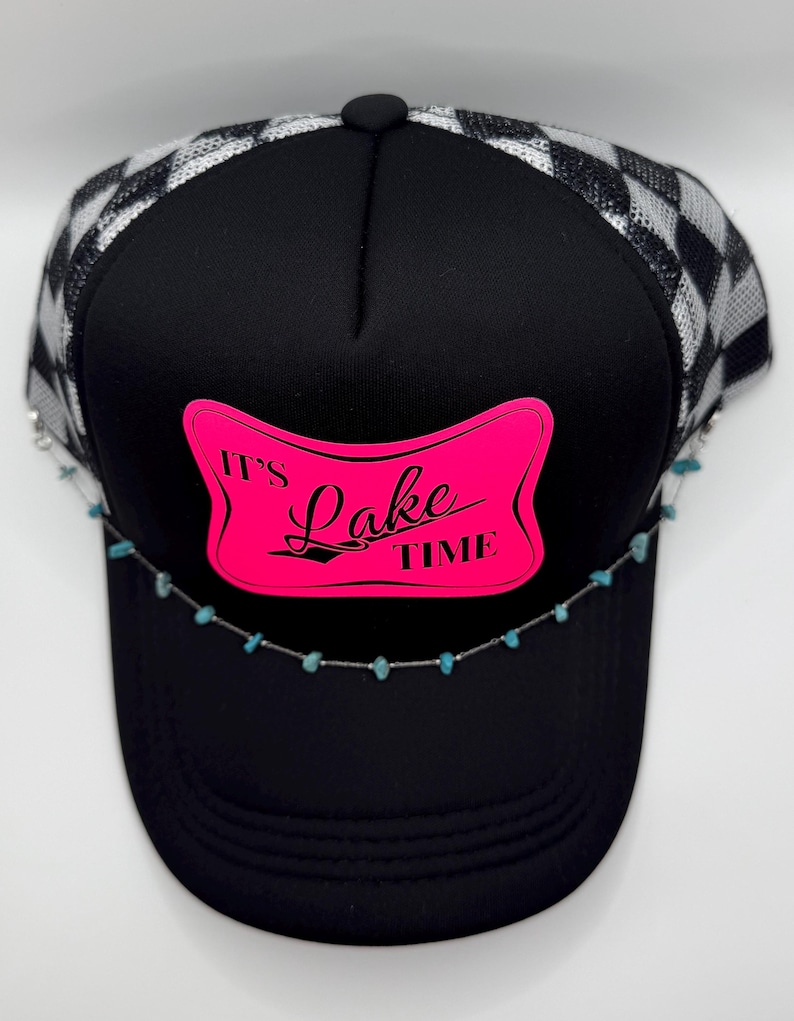 May include: Black trucker hat with a checkered silver and black mesh back. The front features a pink rectangle with the words "It's Lake Time" in black. A turquoise stone chain is attached to the brim.