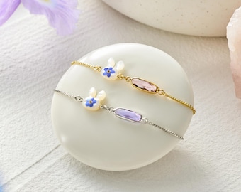 May include: Two delicate bracelets featuring a small white rabbit charm with a blue flower detail and an oval-shaped crystal. One bracelet is gold-toned with a pink crystal, and the other is silver-toned with a light purple crystal.