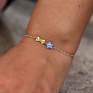 Op de afbeelding: Een goudkleurige kettingarmband met een kleine, platte, botvormige bedel gegraveerd met de naam "Rocky" en een lichtblauwe, vijfbladige bloembedel met een zilverkleurig midden, bevestigd aan een fijne schakelketting om de pols.