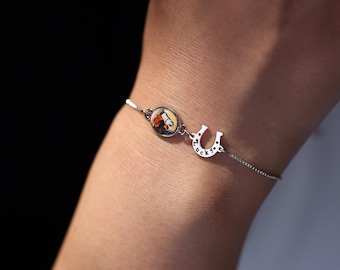 May include: A silver-toned chain bracelet featuring a small oval charm with a floral print and a horseshoe-shaped charm engraved with the word Lucky. The delicate metal chain connects these two distinct decorative elements in a simple design.
