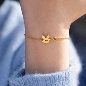 Op de afbeelding: Een goudkleurige kettingarmband met een centraal konijnvormig bedeltje gegraveerd met de naam Nux, vergezeld van een kleine, ronde, donkergekleurde edelsteen. De fijne metalen ketting is aan beide zijden verbonden met de bedel.