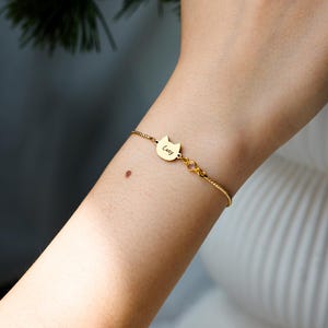 May include: A gold-toned chain bracelet featuring a small, cat-shaped charm engraved with the name Lucy. The bracelet includes a single, small, round, light-colored gemstone accent set in a gold-toned metal link next to the cat charm.