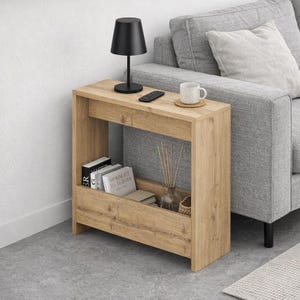 May include: A light brown wooden side table with a black lamp, remote, and a white mug on top. The table has a lower shelf with books and a glass vase with decorative sticks. The table is next to a gray sofa.
