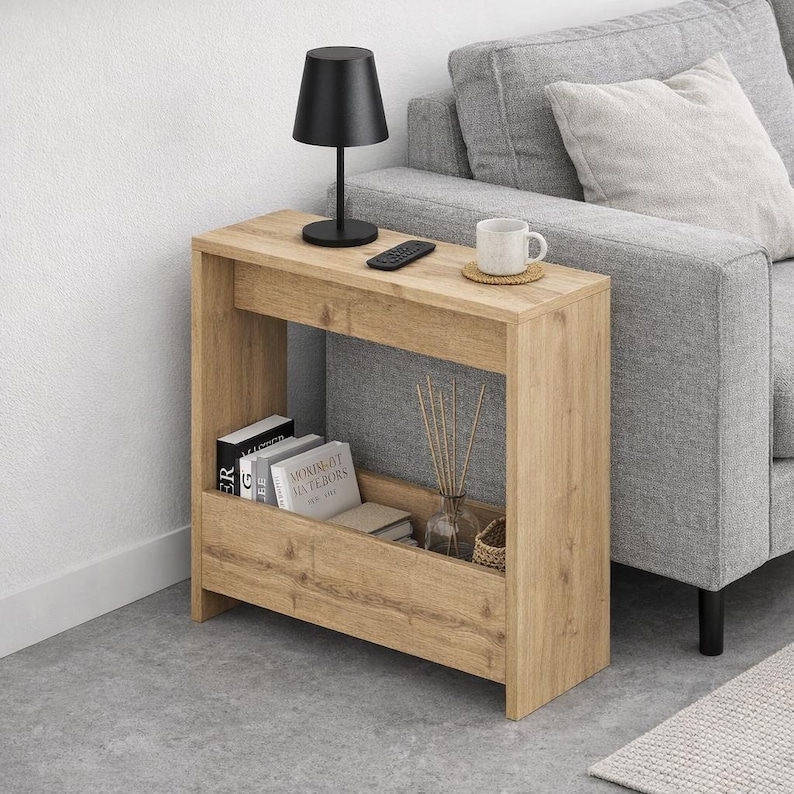 May include: A light brown wooden side table with a black lamp, remote, and a white mug on top. The table has a lower shelf with books and a glass vase with decorative sticks. The table is next to a gray sofa.