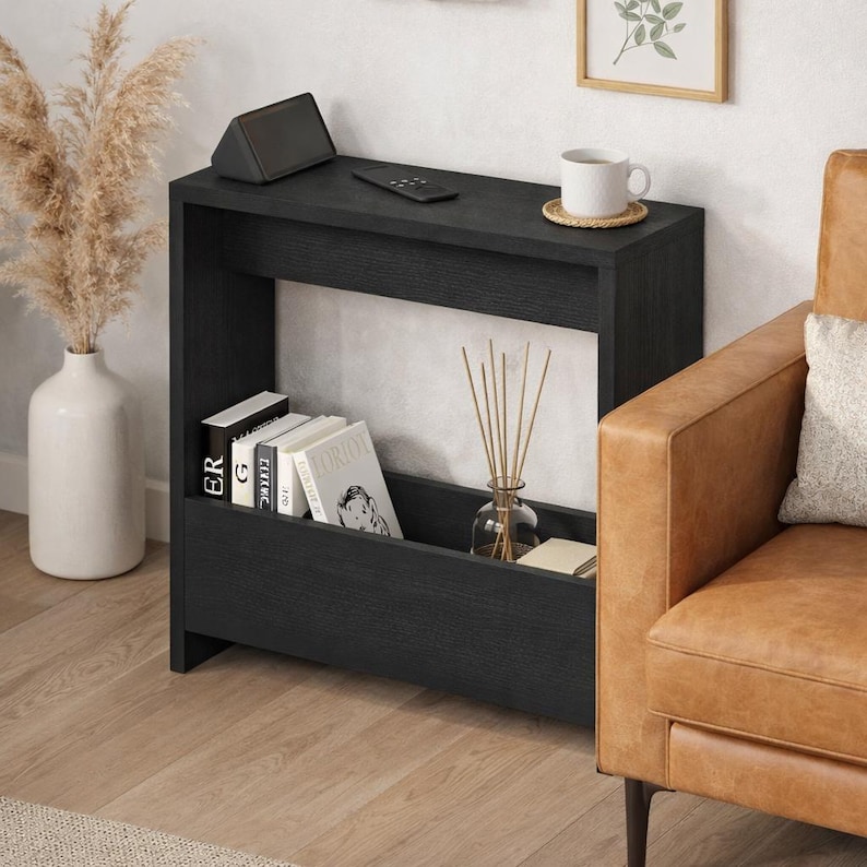 May include: A black wooden side table with a shelf for books and a lower storage area. A small speaker and remote sit on top, with a mug and framed art in the background. A vase with dried plants and a leather armchair are also visible.