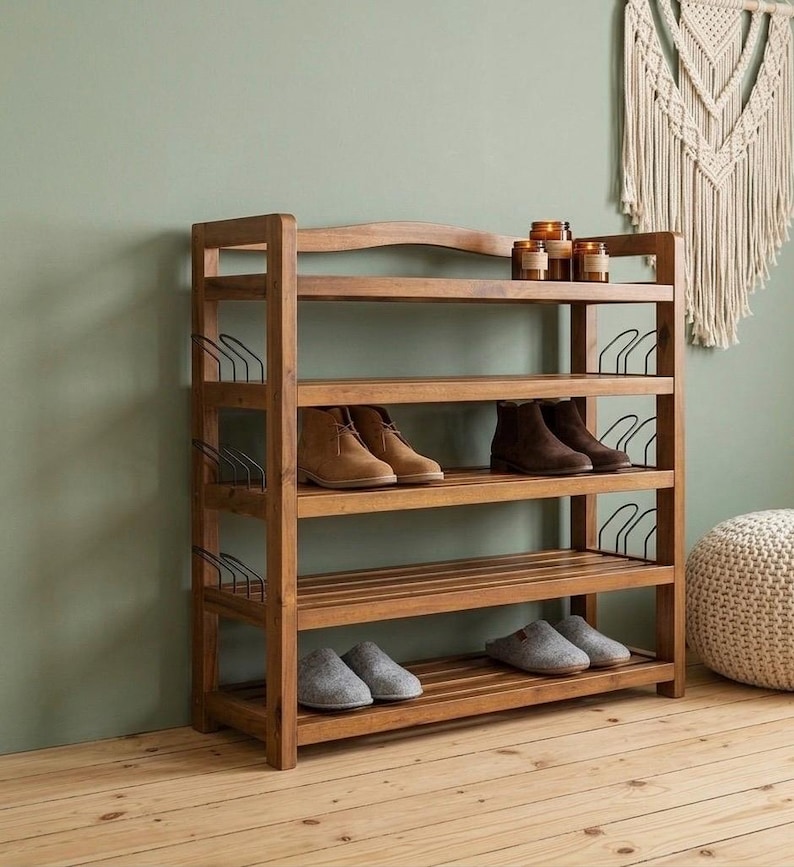 May include: A wooden shoe rack with four shelves, holding various pairs of shoes and slippers. The rack is made of light brown wood and has a simple, functional design. The background features a green wall and a decorative wall hanging.