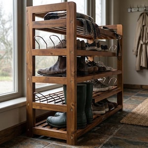 May include: A wooden shoe rack with multiple shelves, holding various shoes, boots, and a folded blanket. The rack is made of dark brown wood and is positioned near a window. The shoes include brown leather boots and green rain boots.