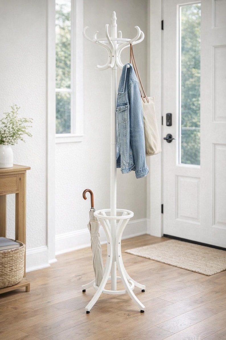 May include: A white coat rack with multiple curved hooks, a central pole, and a circular base. A denim jacket, a beige tote bag, and a light-colored umbrella hang from the rack. The rack is in a bright entryway with a wooden floor.