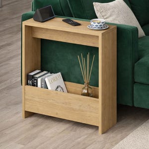 May include: A light oak-colored side table with a built-in magazine rack. The table has a top surface with a small speaker, remote, and a teacup. The rack holds books and a glass diffuser with reeds. The table is next to a green sofa.