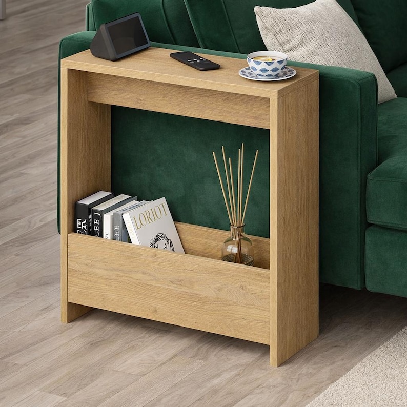 May include: A light oak-colored side table with a built-in magazine rack. The table has a top surface with a small speaker, remote, and a teacup. The rack holds books and a glass diffuser with reeds. The table is next to a green sofa.