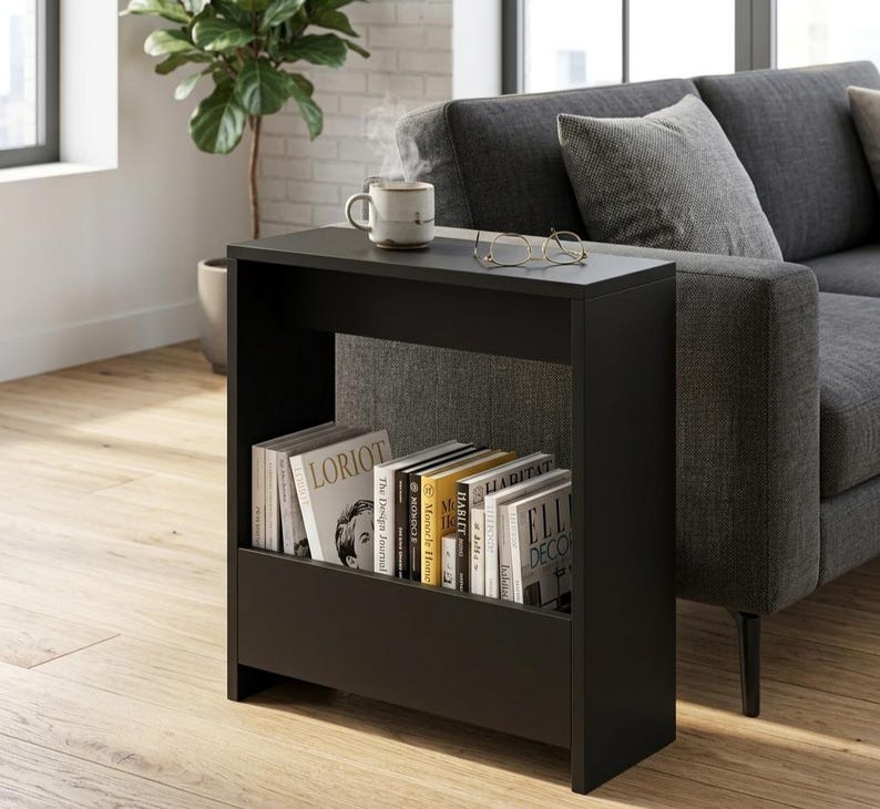 May include: A black side table with a built-in shelf, holding several books. A mug of coffee and eyeglasses rest on the tabletop. The table is next to a gray sofa and a window, with a plant in the background.