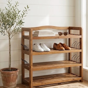 May include: A wooden shoe rack with three shelves, holding white sneakers and brown leather loafers. Sunglasses and books rest on the top shelf. A potted olive tree stands to the left of the rack, against a white wall.