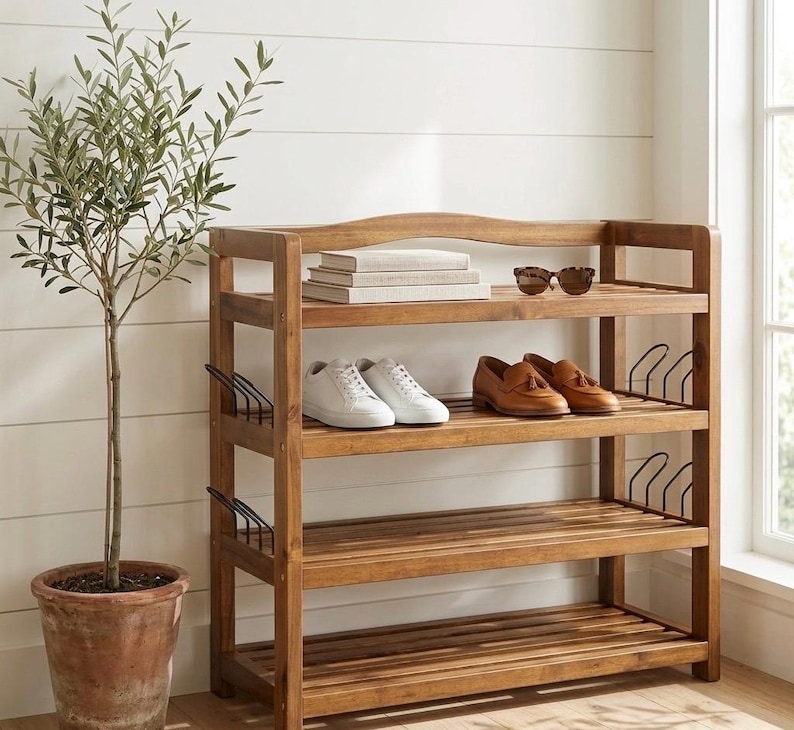 May include: A wooden shoe rack with three shelves, holding white sneakers and brown leather loafers. Sunglasses and books rest on the top shelf. A potted olive tree stands to the left of the rack, against a white wall.