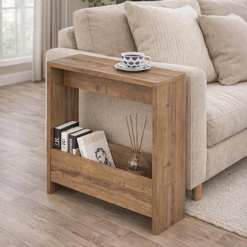 May include: A wooden side table with a built-in shelf, placed next to a beige sofa. The table has a rectangular design with a natural wood finish. A cup and saucer sit on top. Books and a diffuser are in the shelf.