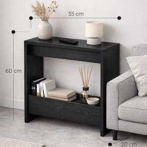 May include: A black console table, 55 cm wide and 60 cm tall, with a lower shelf. It features a vase with dried plants, a lamp, books, and decorative items. The table is placed next to a gray sofa.