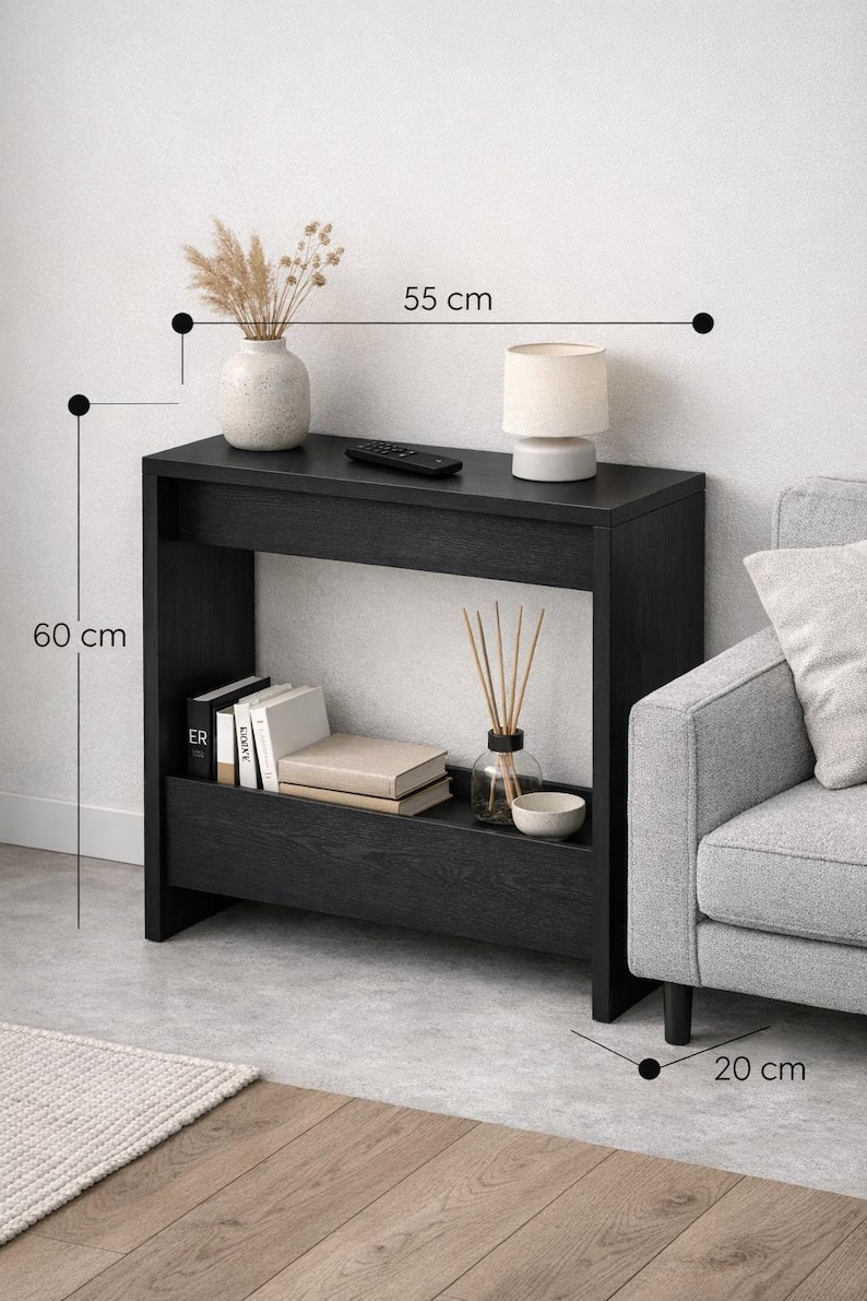 May include: A black console table, 55 cm wide and 60 cm tall, with a lower shelf. It features a vase with dried plants, a lamp, books, and decorative items. The table is placed next to a gray sofa.
