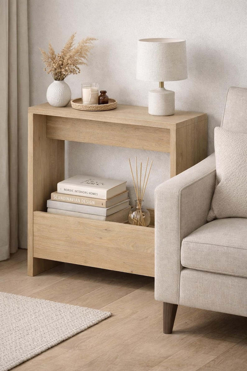 May include: A light oak console table with a lamp, books, and decorative items. The table has a lower shelf with books titled "Nordic Interiors Homes" and "Scandinavian Design". A beige armchair is next to the table.