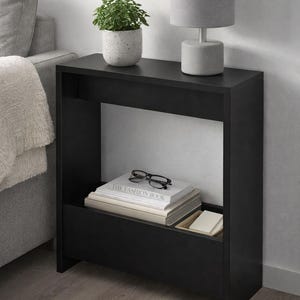 May include: A black side table with a shelf and a lower compartment. A gray lamp and a potted plant sit on top. Books and eyeglasses are in the shelf. The table is next to a gray sofa.