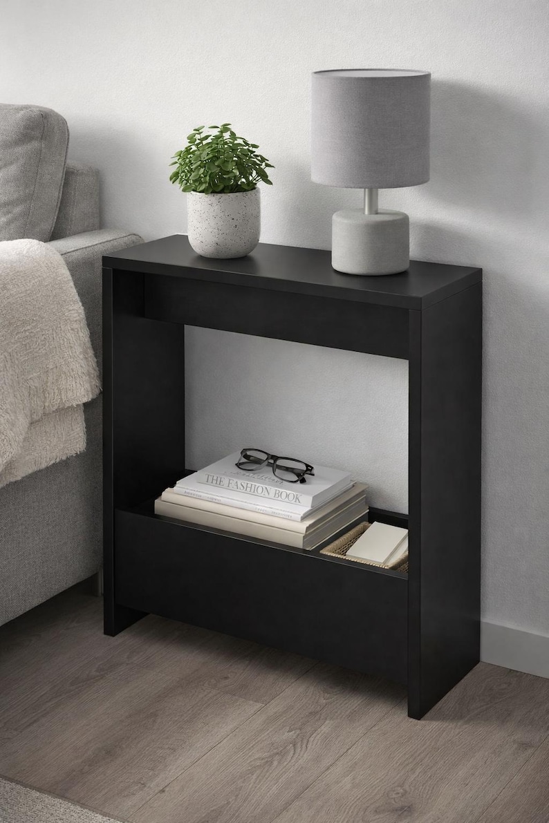 May include: A black side table with a shelf and a lower compartment. A gray lamp and a potted plant sit on top. Books and eyeglasses are in the shelf. The table is next to a gray sofa.
