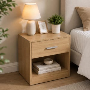Peut inclure: Une table de chevet en chêne clair avec un tiroir et une étagère ouverte. Une lampe blanche avec un abat-jour beige, une petite photo encadrée et une plante en pot sont posées dessus. Le tiroir a une poignée argentée. Des livres et un bol décoratif sont sur l'étagère.