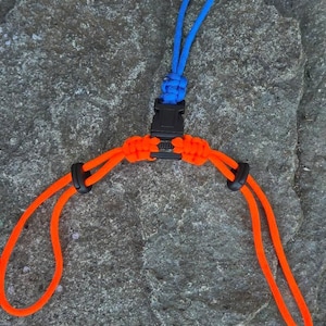 May include: A paracord accessory featuring a central black plastic side-release buckle. A blue cord loop extends from the top, while two orange cord loops extend from the bottom, each secured with a black plastic sliding toggle for adjustment.