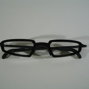 May include: Black plastic framed glasses with a geometric shape. The glasses have a dark lens.