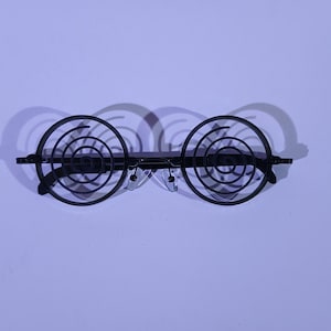 May include: Black spiral-design spectacles with clear nose pads. The lenses are circular with a spiral pattern. The spectacles are set against a light purple background, casting a shadow.