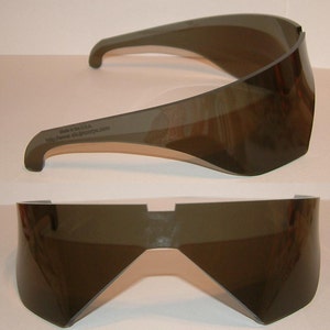 May include: A pair of dark gray plastic sunglasses with a futuristic design. The sunglasses are made in the USA and the website address is printed on the side: http://www.skigoggles.com