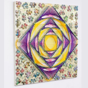 May include: A square, handmade string art piece with a geometric design. The artwork features a central yellow and purple diamond pattern, surrounded by a background of puzzle pieces. The piece is mounted on a white surface.