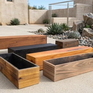 May include: A collection of rectangular wooden planters and blocks in various shades of brown and black. The planters are open-topped, and the blocks are solid. The wood grain is visible, and the items are arranged on a concrete surface.