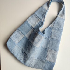 Könnte beinhalten: Eine Umhängetasche im Hobo-Stil, gefertigt aus einem Patchwork aus hellblauen und verwaschenen Denim-Quadraten. Die Tasche verfügt über einen integrierten Schulterriemen und eine weiche, unstrukturierte Form mit sichtbaren Nähten an den Stoffteilen.