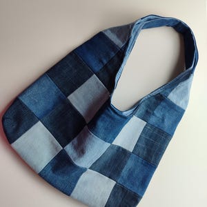 Könnte beinhalten: Eine Umhängetasche im Hobo-Stil, gefertigt aus einem Patchwork aus quadratischen Denim-Stoffstücken in verschiedenen Blautönen, die von hellem Himmelblau bis zu tiefem Indigo reichen. Die Tasche verfügt über einen integrierten Schulterriemen.