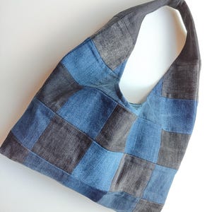 Könnte beinhalten: Eine Hobo-Tasche aus einem Patchwork aus quadratischen Denim-Stoffstücken in abwechselnden Farbtönen von Mittelblau und Dunkelanthrazit. Die Tasche verfügt über einen einzelnen integrierten Schulterriemen und eine lockere, unstrukturierte Form.