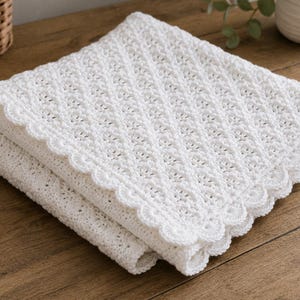 May include: A white crocheted blanket featuring a repeating diamond pattern throughout the centre and a scalloped edge border. The textile is folded into a square shape, showcasing the intricate yarn work and the consistent white colour.