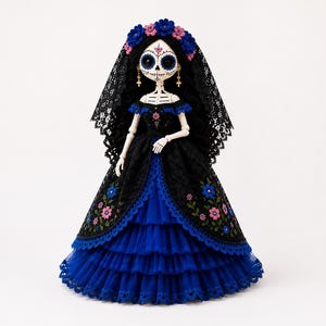 Puede incluir: Una muñeca esquelética con rostro blanco, cuencas oculares negras y detalles florales, que lleva un velo de encaje negro con flores azules y rosas. La muñeca viste un vestido de capas negro y azul con bordados florales y pendientes de cruz.