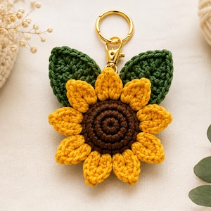 Op de afbeelding: Een gehaakte zonnebloem sleutelhanger met een donkerbruin hart, felgele bloemblaadjes en twee donkergroene bladeren. De bloem is bevestigd aan een goudkleurige metalen karabijnhaak en een ronde sleutelring als decoratief accessoire.