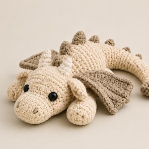 May include: A crocheted dragon toy in cream and light brown yarn. The creature features two black plastic eyes, small horns, textured wings, and a long tail with a spade-shaped tip. It has soft pink blush details on its cheeks and a series of brown spikes along its back.