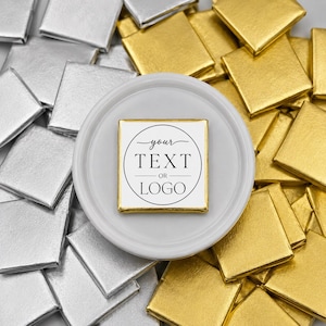 May include: A square chocolate piece with a white wrapper featuring the black text 'your TEXT OR LOGO' inside a circle, centred on a white circular plate surrounded by a pile of numerous square chocolates wrapped in shiny gold and silver foil.