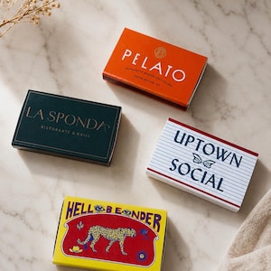 May include: Four rectangular matchboxes with distinct designs. One is orange with white text reading PELATO, one is dark green with gold text reading LA SPONDA, one is white with blue lined paper and text UPTOWN SOCIAL, and one is yellow with a red leopard graphic.