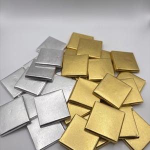 May include: A collection of square, metallic tiles in silver and gold. The tiles are arranged in a loose pile, with the silver tiles on the left and the gold tiles on the right. The tiles have a slightly textured surface and a reflective sheen.