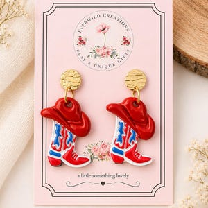 Può includere: Un paio di orecchini pendenti a forma di stivali da cowboy rossi con accenti blu e bianchi, sormontati da un cappello da cowboy rosso. Gli orecchini pendono da perni circolari dorati testurizzati su un cartoncino rosa Everwild Creations.