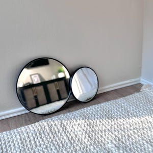 May include: Two round mirrors with black frames, one large and one small, are positioned against a light-colored wall. The mirrors reflect the room's interior, including a cabinet and window light. The mirrors are placed on a wooden floor next to a white rug.