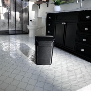 May include: A small, black, rectangular trash can with a hinged lid. The trash can is in a bathroom setting with a white tile floor and a black vanity.