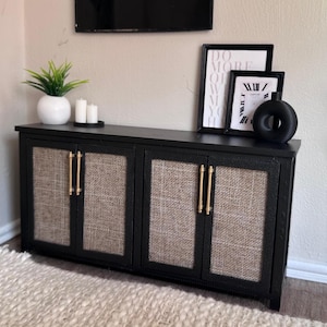 Puede incluir: Un mueble negro en miniatura con cuatro puertas, cada una con un panel beige tejido y tiradores dorados. El mueble está coronado con una maceta blanca, velas y obras de arte enmarcadas. Un objeto decorativo negro se encuentra en el mueble.
