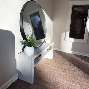 May include: A miniature white console table with a round black framed mirror. The table holds a small white vase with greenery and a stack of books. The room has a wood floor and a window.