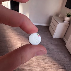 Puede incluir: Un detector de humo blanco en miniatura con luces indicadoras rojas y verdes. El pequeño dispositivo redondo tiene un diseño estriado y se sostiene entre los dedos. El fondo muestra una habitación en miniatura con una cómoda y una planta.