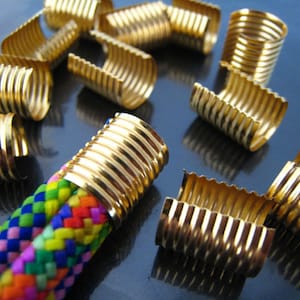 Puede incluir: Primer plano de terminales de cordón de metal dorado. Los terminales de cordón son acanalados y tienen un acabado brillante. Los terminales de cordón están dispersos sobre una superficie azul oscuro. Un cordón trenzado de colores está enhebrado a través de uno de los terminales de cordón.