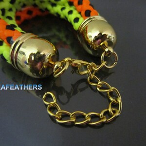 Puede incluir: Un brazalete trenzado negro, naranja y verde con tapas de extremo doradas y un cierre de cadena dorada.